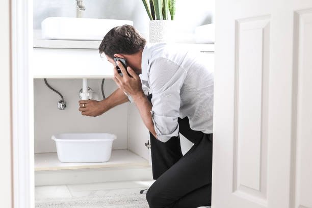 Sad Young Man Calling Plumber In Front Of Water Leaking From Sink Pipe